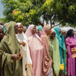 A group of women in colorful hijabs gathering under a tree on a sunny day.