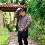 A stylish cowboy tips his hat while posing in a lush garden setting.